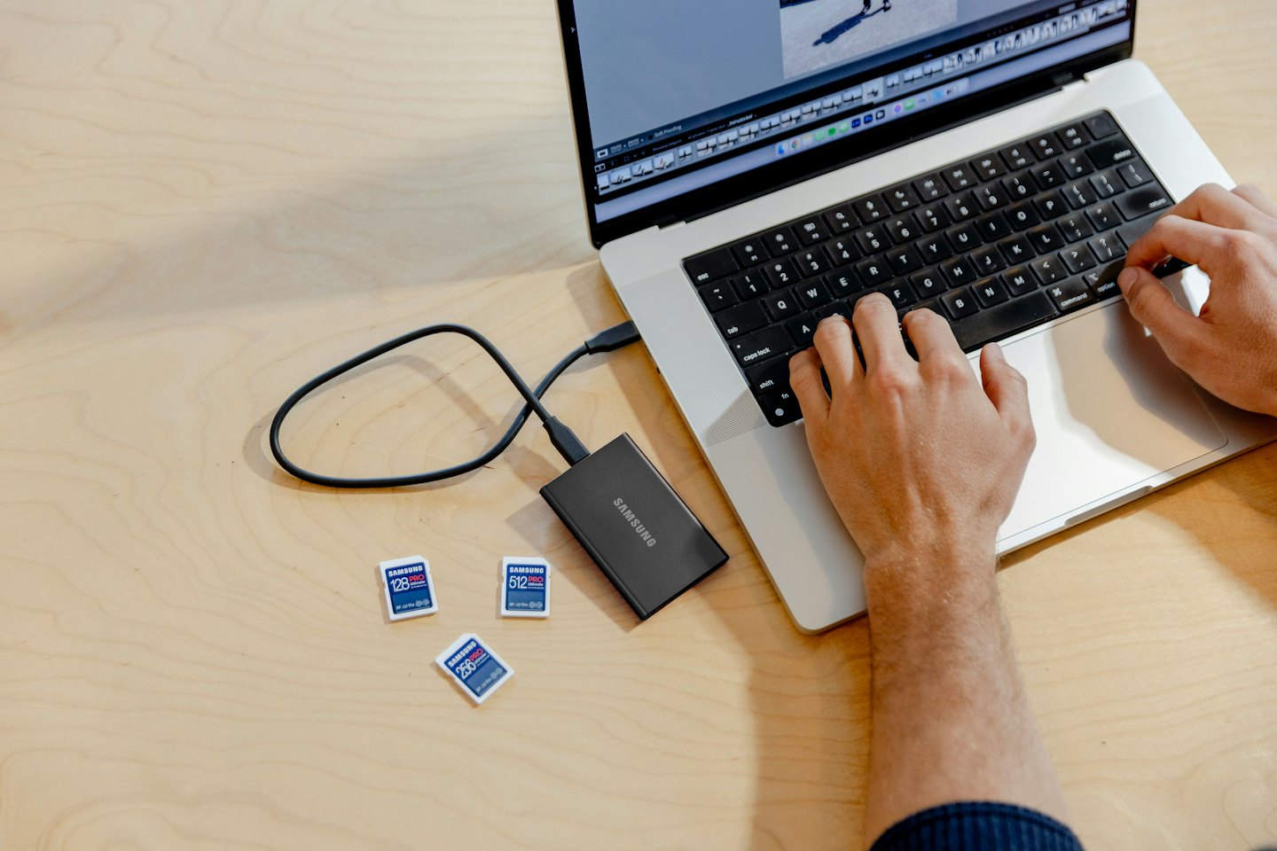 Hands typing on a laptop keyboard with an external hard drive and microSD cards.